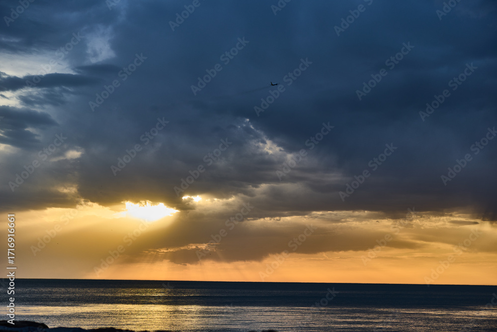 Obraz premium dramatic sunset ocean horizon with sun rays breaking through dark storm clouds over calm sea water and distant airplane silhouette