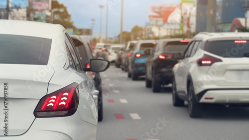 Rear side of white car and turn on brake light. Traffic is congested and there are long queues in the city. Blurred image of traffic jam with town under the blue sky.