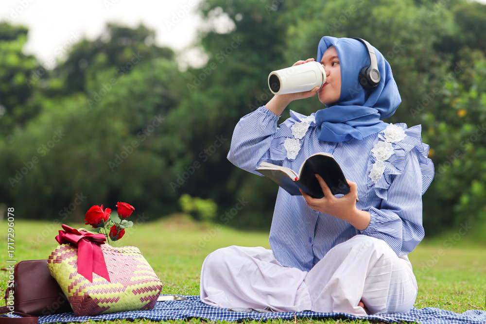 Φωτογραφία Beautiful Asian Muslim woman sitting on the ground in the park and drinking a cup of tea in a thermos or personal glass while listening to music in the afternoon