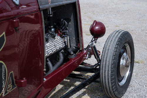 A close-up of the exposed V8 engine and front wheel of a classic American hot rod. The detailed shot highlights the mechanical parts and chrome suspension