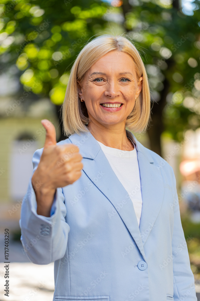 Φωτογραφία Happy smiling mature Caucasian blonde businesswoman lady female woman in formal suit looking camera and showing thumb up hand, positive sign gesture like recommend well-done outdoors city town street