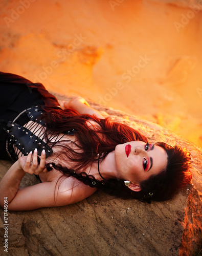 A young woman dressed in leather clothes lies on a stone against the background of a fiery backlight.
