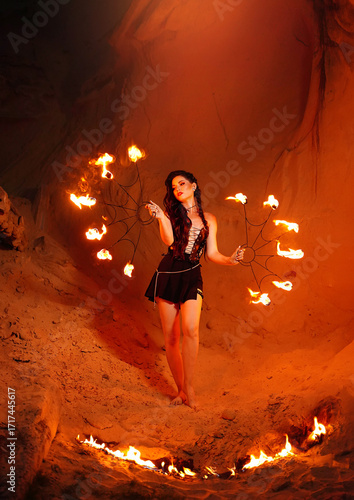 A young woman dressed in leather clothes stands on the sand and a flame of fire next to her. Fire show in the desert.