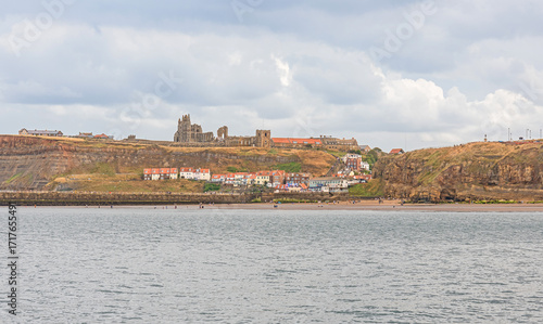 A seaside panorama with the ruins of an abbey on a hill and houses beneath.