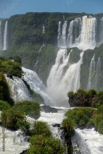 Iguazu Falls, showcasing its multiple, powerful cascades and a vibrant rainbow arcing through the mist. The shot captures the vast scale and stunning natural beauty of the site, making it ideal for to