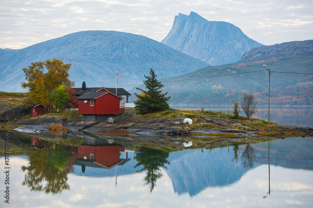 Fototapeta premium Red cabins by a fjord near Tromsø, Norway. Autumn trees and dramatic mountains reflected in calm water, creating a peaceful Nordic landscape.