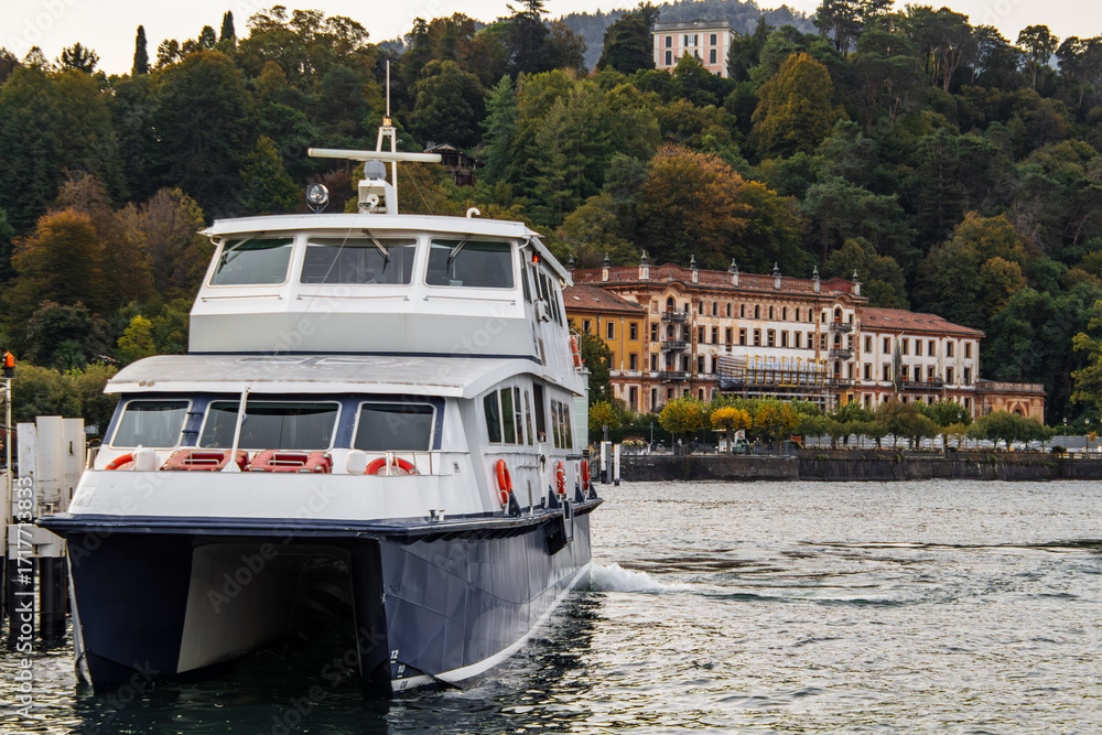 Naklejka premium Modern catamaran ferry departs Lake Como, past historic grand buildings and lush autumn trees on the tranquil Italian shore. Picturesque landscape, classic charm, calm waters.