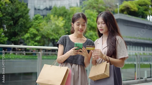 Two asian beautiful girl friends standing on the street with their purchases in shopping bags. The woman making payment online using her cellphone and credit card. Shopaholism, easy payment