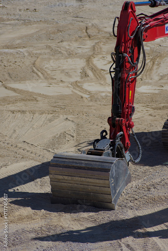 The powerful arm and digging bucket of a red excavator rest on a sandy construction site with tire tracks visible in the background.