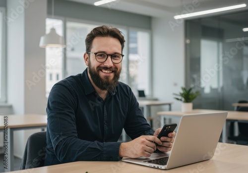Smiling bearded man with glasses holding a smartphone while sitting at a desk with a laptop in a modern office environment