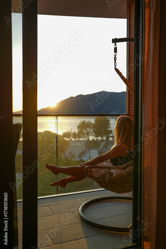 Canvas Print Young blonde woman relaxing in a hanging chair on a balcony, gazing at the sunset over the sea and mountains