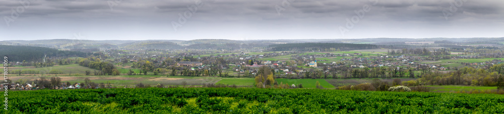 Fototapeta premium A panoramic view of Mliyiv village, Cherkasy region, Ukraine, in the Vilshanka River valley, during spring late April. The photo shows houses and buildings surrounded by green fields and rolling hills