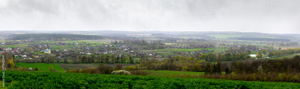 Fototapeta premium A panoramic view of Mliyiv village, Cherkasy region, Ukraine, in the Vilshanka River valley, during spring late April. The photo shows houses and buildings surrounded by green fields and rolling hills
