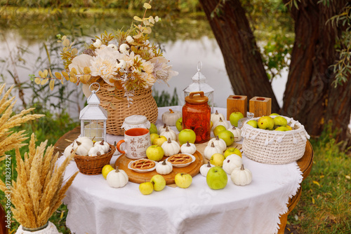 Rustic autumn table setting is arranged outdoors, next to a calm river. The round table is covered with a white cloth and is laden with seasonal harvest decor. 