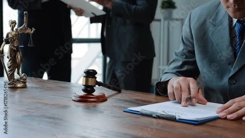 Lawyer in suit and tie sitting at desk with gavel and statue of Lady Justice, reviewing and signing a contract while another person stands in background