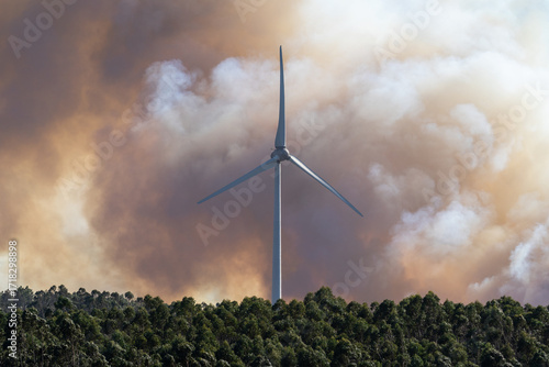 A dramatic wildfire burns near a windmill under a sky filled with thick clouds. The contrast between renewable energy and destructive flames highlights themes of climate change, environmental crisis, 