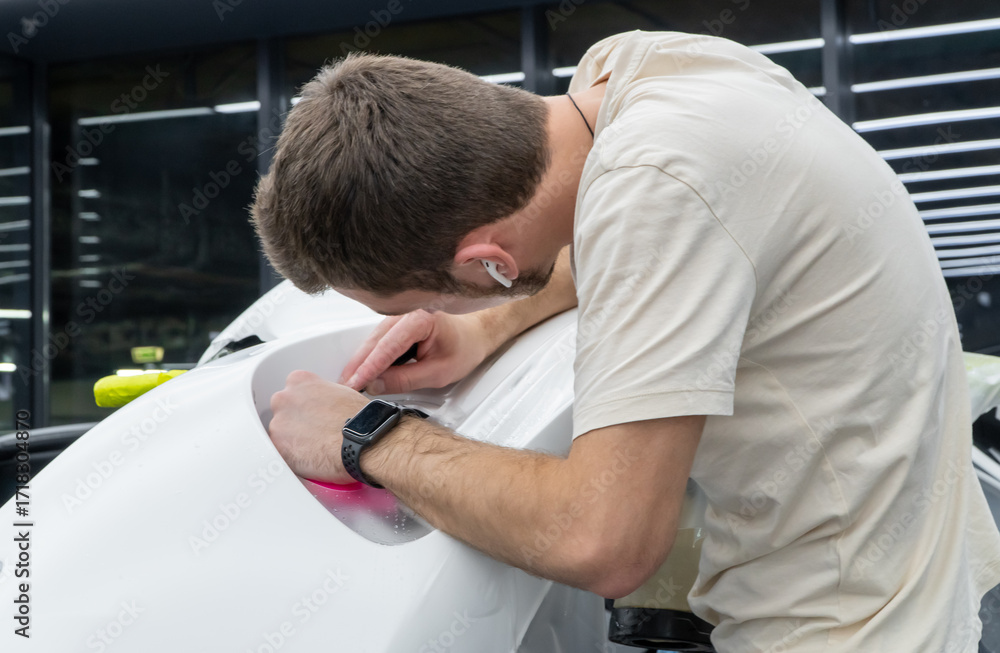 Obraz premium A car wrapping specialist applies a polyurethane film to the car. Detailer smoothes PPF. Car wrapping, detailing, tuning and styling. PPF protective film against chips and scratches. Selective focus.