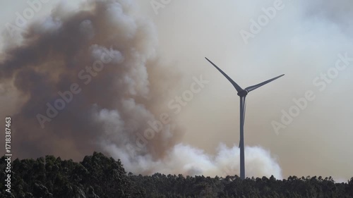A dramatic wildfire burns near a windmill under a sky filled with thick clouds. The contrast between renewable energy and destructive flames highlights themes of climate change, environmental crisis
