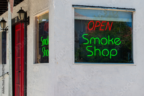 Two large glass windows in a tobacco shop with a red open sign and a green smoke shop sign. The vintage porcelain neon signs hang from corner windows. There's a red door with multiple glass windows.