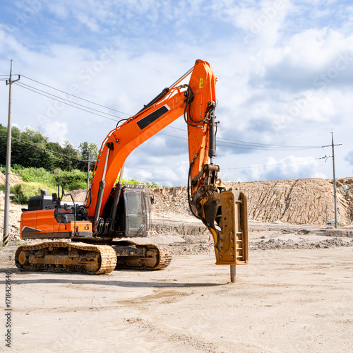 Yellow excavator with a hydraulic breaker attachment working at a construction site on a sunny day. Heavy machinery used for demolition, rock breaking, and ground preparation. Suitable for constructio