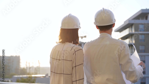 Billede på lærred Man and woman architects constructor engineers wearing safety glasses and hardhats, are examining blueprints at a construction site during a sunset, back view