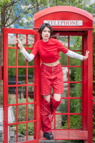 A pretty young woman in a modern red outfit is inside a red public phone booth, holding a vintage pay phone. A cute teenage girl expresses a variety of emotions while talking.