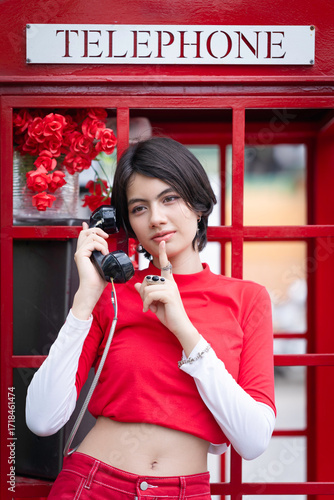 A pretty young woman in a modern red outfit is inside a red public phone booth, holding a vintage pay phone. A cute teenage girl expresses a variety of emotions while talking.