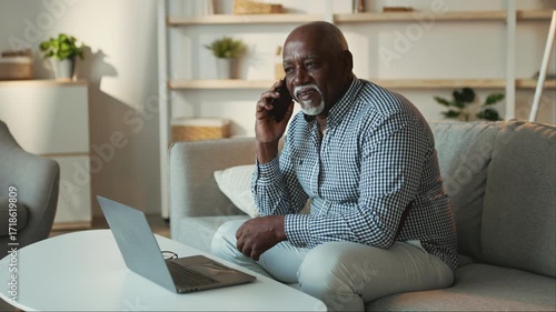 An elderly African American man sits comfortably on a couch, talking on the phone while working on a laptop. He appears focused and relaxed in a cozy living room setting.