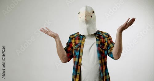 A man wearing a bird mask shrugs his shoulders. He is confused and unsure. He gestures with his hands, palms up. He is wearing a plaid shirt. The background is white.