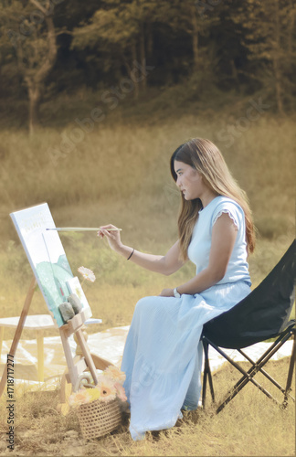 Side view of a beautiful asian woman sit on chair and drawing nature, landscape view painting on canvas background. Photograph of young female painter creating art in an outdoor setting.