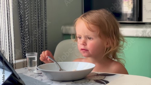 Little girl sits at the table eating porridge while watching cartoons on a tablet. Modern childhood with gadgets