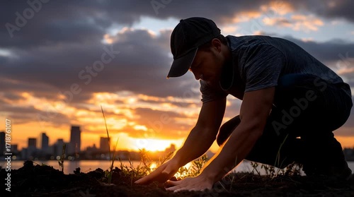A man in a cap tending to grass at sunset with city skyline in the background and cloudy sky above