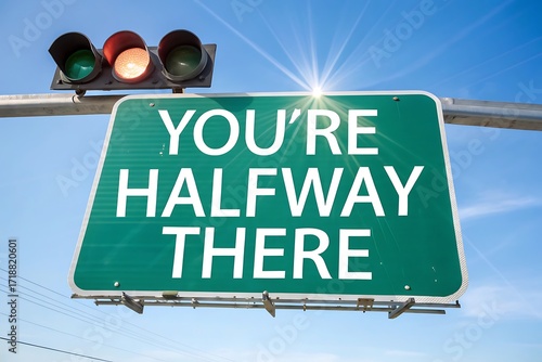 Bright green highway sign reads you're halfway there against blue sky traffic light