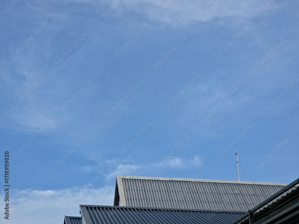 Fototapeta premium a bright blue sky with thin, wispy cirrus clouds above the sloped, corrugated metal roofs of several buildings.