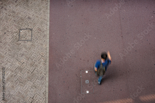 Top-down shot captures motion of pedestrian with backpack walking with quiet intent along a red asphalt path beside brick-paved sidewalk, highlighting urban textures, motion, and everyday city life.
