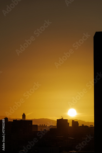 Vibrant sunrise paints morning sky rich golden hue, softly blending colors as brilliant globe hovers over faint hazy line of distant hills, contrasting foreground's densely packed silhouetted city.