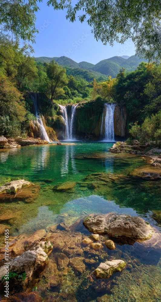 Obraz premium Serene waterfall cascading into a crystal-clear pool, surrounded by lush greenery and mountains under a sunny sky, viewed from a low angle