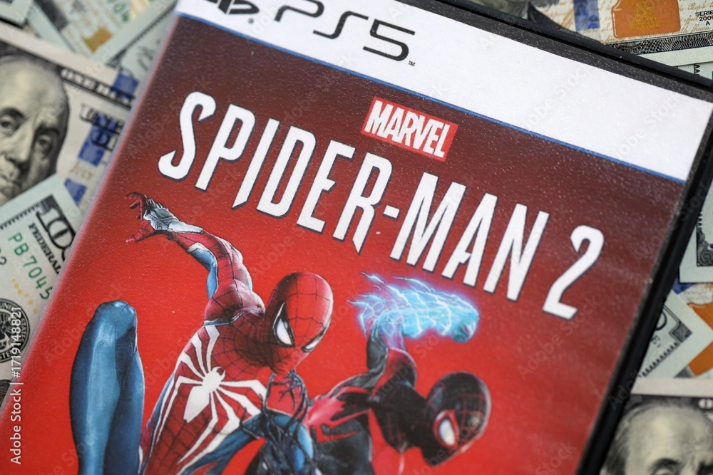 Naklejka premium KYIV, UKRAINE - SEPTEMBER 8, 2025 Marvels Spider-Man 2 videogame lies on big amount of hundred dollar bills