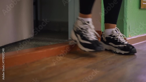 Close view of legs in white and black sneakers stepping out through doorway, casual exit captured with focus on shoes and floor details, indoor background showing textured wall and polished wooden