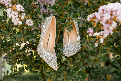 Sparkling silver high heels with rhinestone embellishments displayed on a blooming shrub. Elegant bridal shoes captured in a stylish outdoor wedding detail shot. Women's festive shoes