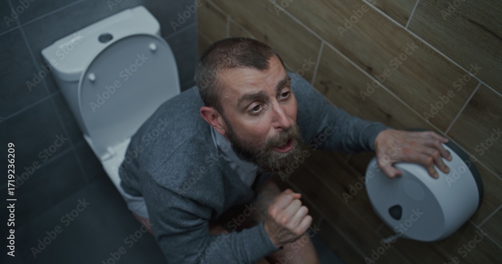 Obraz premium Businessman with Tense Facial Expression Suffering from Diarrhea and Indigestion, Sitting on the Toilet. Man Experiencing Upset Stomach, Digestive Discomfort in Office Restroom During Work. High Angle