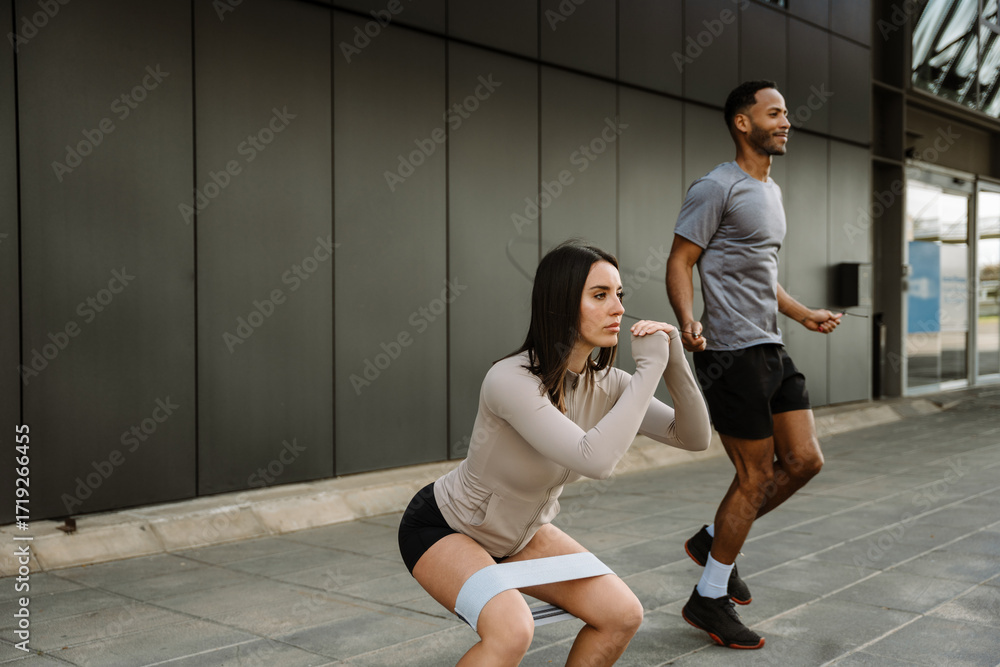 Fototapeta premium A female athlete squats with a rubber band on her hips while a male athlete jumps rope next to her