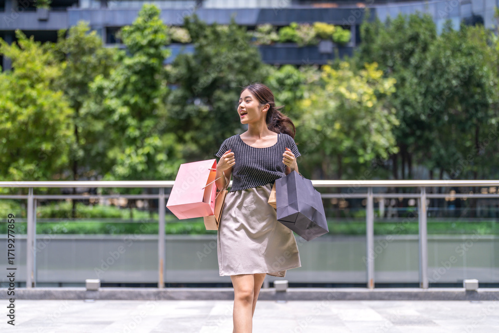 Fototapeta premium Happy asian woman walking outdoors with shopping bags at mall, joyful shopping fashion, consumer happiness, weekend activity and celebrating purchase, sale promotion, cashback reward after shopping