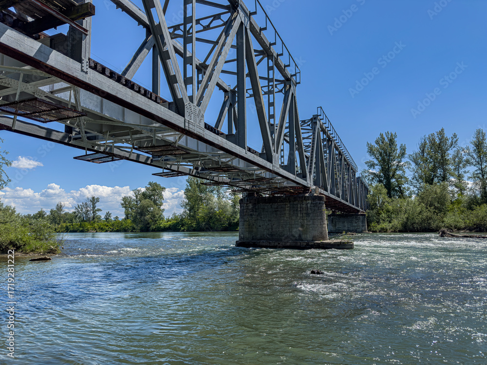 Obraz premium A steel truss bridge spans across a flowing river surrounded by lush greenery under a bright summer sky. The Tisza River. Transcarpathia, Ukraine.