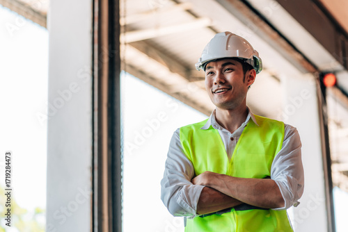 A male warehouse manager in a white shirt, a white hard hat, and a neon green reflective vest stands with his arms crossed. He is looking camera.a sense of professional supervision and order.