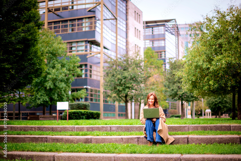 Fototapeta premium Full length of a confident woman with laptop sitting at city park in background with office building and working