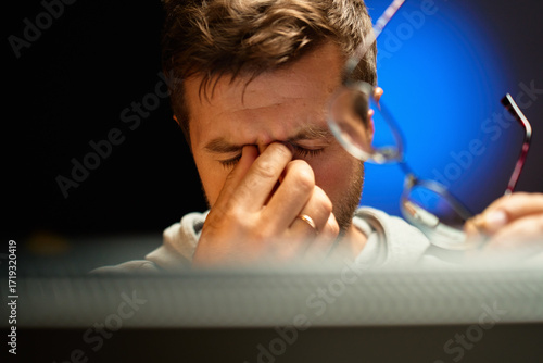 Close-up of tired man rubbing his eyes and holding glasses while sitting at computer. Person has fatigue and headache from overwork and eye strain