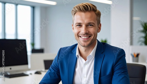 a smiling thirty blonde man with short hair wearing a blue jacket and a white shirt sitting at the desk in the office