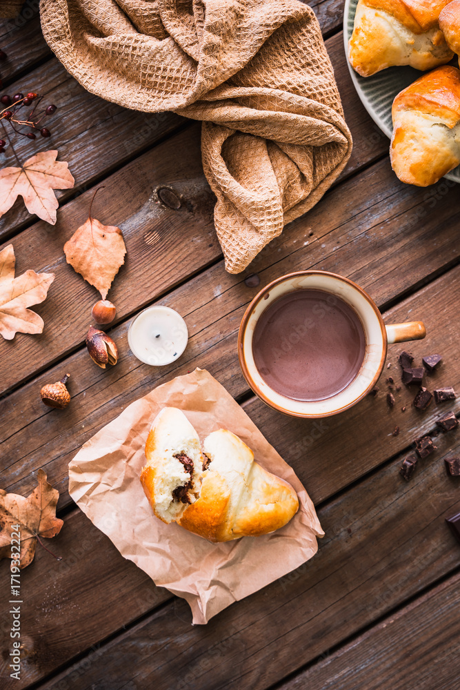 Fototapeta premium Ein gebackenes Schokoladekipferl auf einem Backpapier und eine Tasse Kakao auf einem rustikalen Holz Tisch. Drausicht, draußen, Herbst.