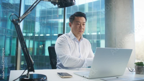 Focused Asian man in elegant shirt putting on headphones while opening laptop in daylight office. Getting ready for virtual meeting or online call. Preparing to join conference or start webinar.